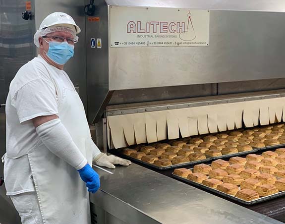 Baker Boy worker standing next to a conveyor belt with freshly baked square croissants.